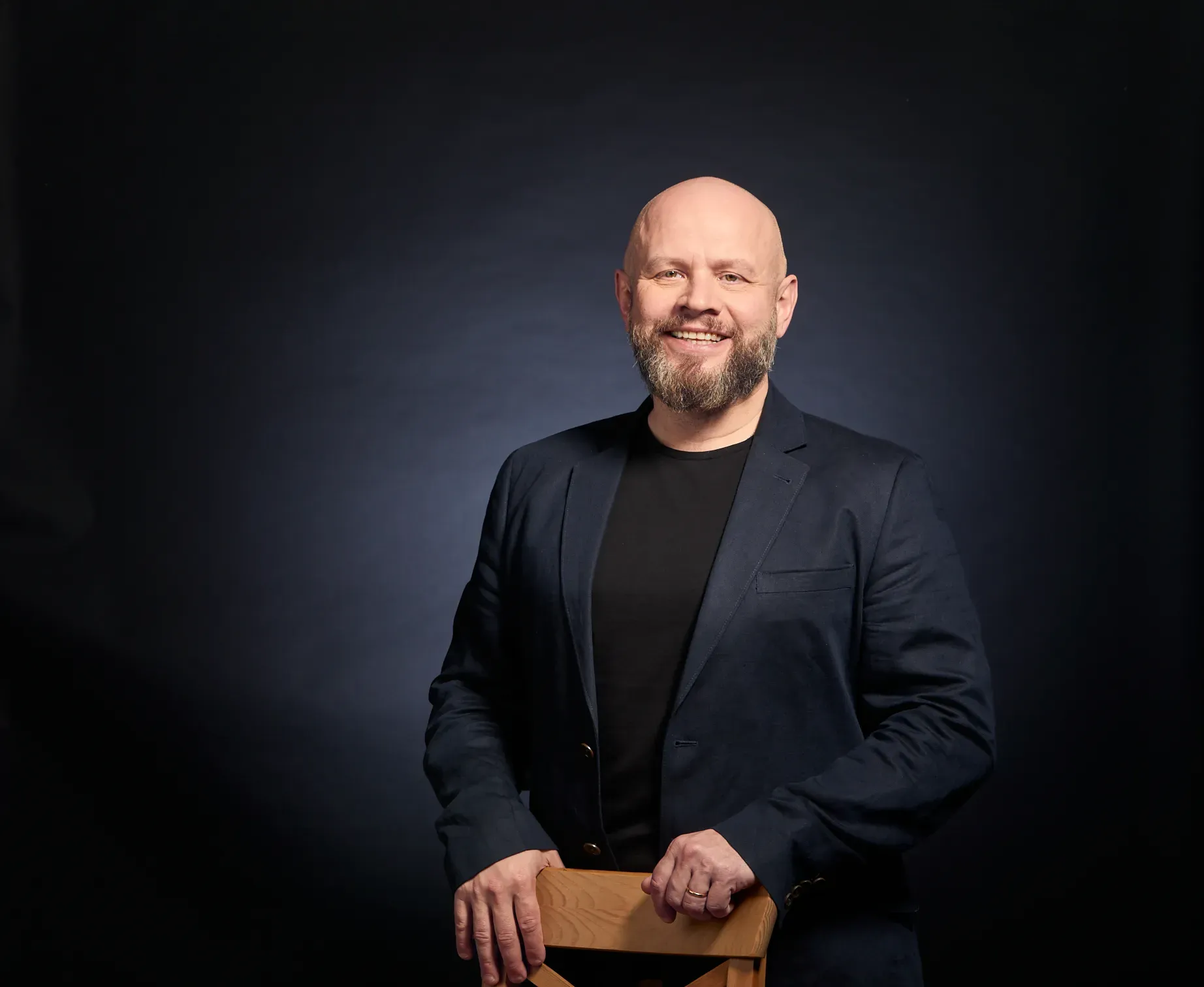 Alexander Golev standing behind a wooden chair, smiling, in a dark blazer against a dark studio background